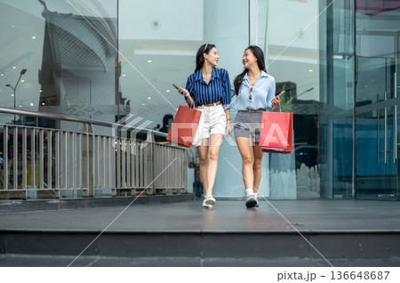 Pretty asian woman and friend holding phone carrying shopping bags while walking in front of a mall. Pretty asian woman and friend holding phone carrying shopping bags while walking in front of a mall. 136648687