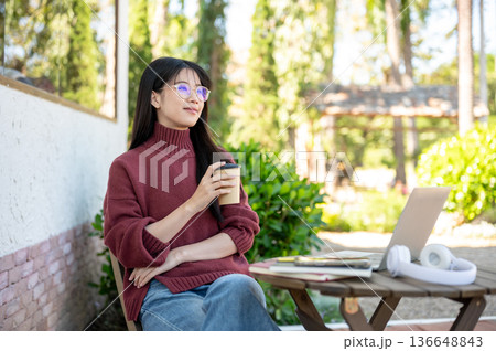 Glasses asian woman in sweater holding coffee aside laptop as sitting at wooden table in garden cafe Glasses asian woman in sweater holding coffee aside laptop as sitting at wooden table in garden cafe 136648843