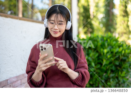 Glasses asian woman listening to music in headphones looking at phone while sitting in garden cafe. Glasses asian woman listening to music in headphones looking at phone while sitting in garden cafe. 136648850