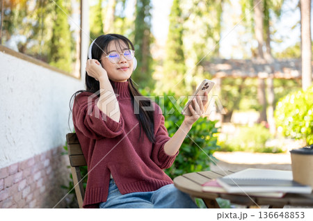 Glasses woman closing eyes listening to music and holding phone sitting at wood table in garden cafe Glasses woman closing eyes listening to music and holding phone sitting at wood table in garden cafe 136648853