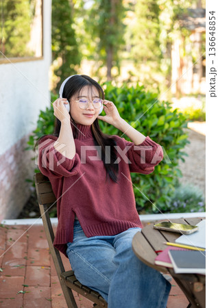 Glasses woman closing eyes listening to music in headphones sitting at wooden table in garden cafe. 136648854