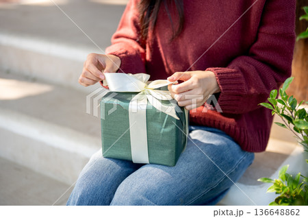 Close up of woman in sweater removing or tying a ribbon on present gift box while sitting on stairs. Close up of woman in sweater removing or tying a ribbon on present gift box while sitting on stairs. 136648862