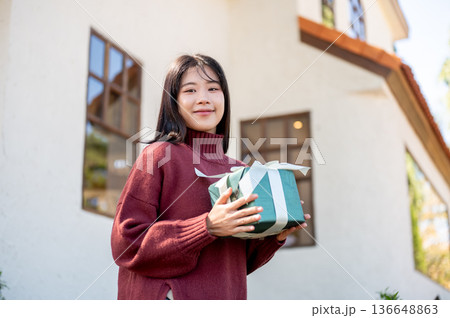 Pretty asian woman in sweater holding present gift box while standing aside a home or cafe building. 136648863