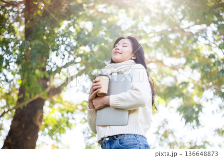 Asian woman in scarf holding coffee hugging laptop standing or walking under a tree in garden park. Asian woman in scarf holding coffee hugging laptop standing or walking under a tree in garden park. 136648873