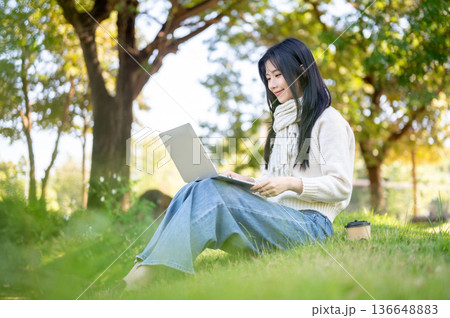 Asian woman in scarf looking using laptop on knees while sitting on grass in garden or nature park. 136648883