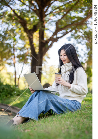 Asian woman in scarf holding coffee looking at laptop while sitting on grass in garden nature park. 136648884