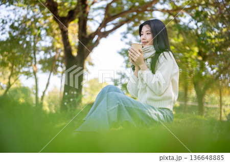 Asian woman in scarf holding hot coffee in both hands while sitting on grass in garden nature park. 136648885