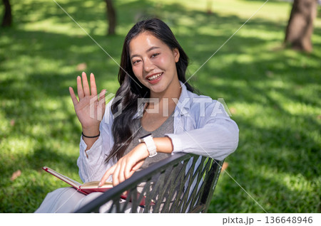 Asian woman holding book and waving a hand while sitting on bench over green grass in garden park. 136648946