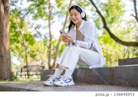 Asian woman looking at phone listening to music in headphone sitting on cement stairs in garden park 136648953