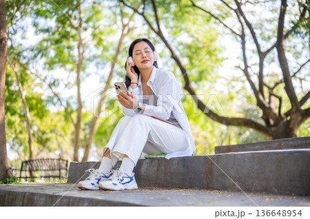 Asian woman closing eyes listening to music in headphones and sitting on cement stair in garden park 136648954