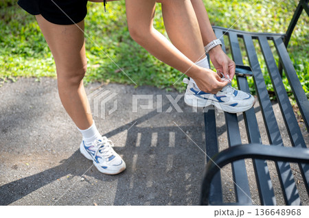 Close up of woman tying sneaker's shoelace on bench before exercise and standing on pavement in park 136648968