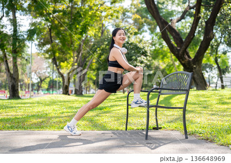 Asian woman in sportswear stretching leg on bench before exercise while standing on pavement in park Asian woman in sportswear stretching leg on bench before exercise while standing on pavement in park 136648969