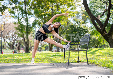 Asian woman in sportswear standing stretching muscles before exercise on bench over pavement in park Asian woman in sportswear standing stretching muscles before exercise on bench over pavement in park 136648970