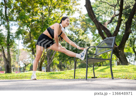 Asian woman in sportswear standing stretching muscle before exercise on bench over pavement in park. 136648971
