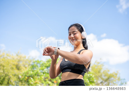 Asian woman in sportswear listening to music in headphones looking at watch while exercise in park. Asian woman in sportswear listening to music in headphones looking at watch while exercise in park. 136648983