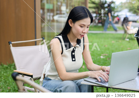 Asian woman student looking and typing on laptop while sitting at table on grass in an outdoors cafe Asian woman student looking and typing on laptop while sitting at table on grass in an outdoors cafe 136649025