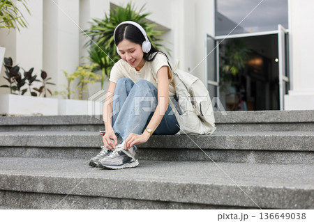 Asian woman student listening to music in headphone tying shoelace as sitting in front of a building Asian woman student listening to music in headphone tying shoelace as sitting in front of a building 136649038