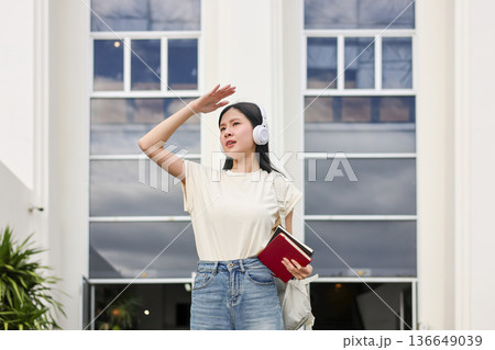 Asian woman student holding book using hand covering eye from sunlight standing in front of building 136649039