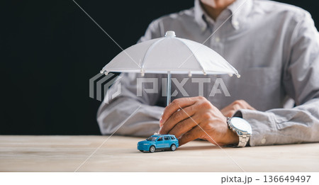 Insurance concept highlights vehicle protection, hand holds small umbrella shielding blue auto from accident risk, driver buys policy for security and future finance management plan Insurance concept highlights vehicle protection, hand holds small umbrella shielding blue auto from accident risk, driver buys policy for security and future finance management plan 136649497