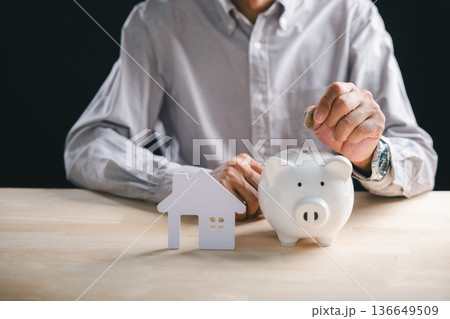 Estate property investment concept, person carefully saving money for future home ownership, placing coin in white piggy bank, showing financial planning for real house dream 136649509
