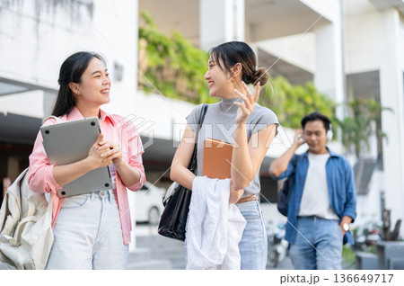 Asian woman student holding laptop talking to a friend holding book standing walking in university Asian woman student holding laptop talking to a friend holding book standing walking in university 136649717