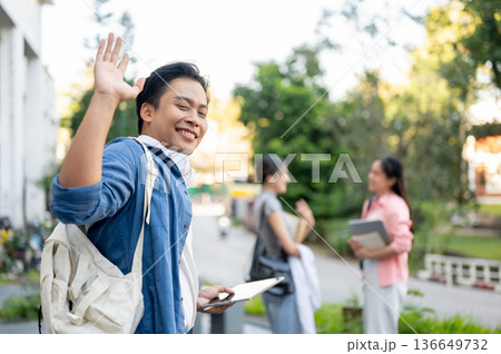 Happy asian man student holding tablet waving a hand while walking to woman friends in university Happy asian man student holding tablet waving a hand while walking to woman friends in university 136649732