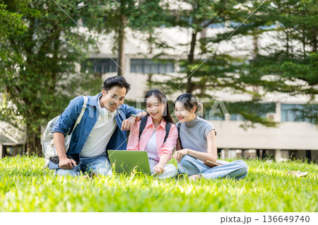 Asian man woman student friends looking at laptop while sitting on grass in university's garden park Asian man woman student friends looking at laptop while sitting on grass in university's garden park 136649740