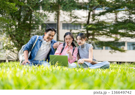 Smiling asian man woman student friends looking at laptop and sitting on grass in university's park Smiling asian man woman student friends looking at laptop and sitting on grass in university's park 136649742