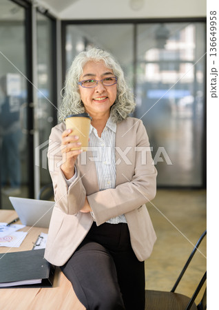 Old asian businesswoman holding coffee cup while standing or sitting leaning on table in office. 136649958
