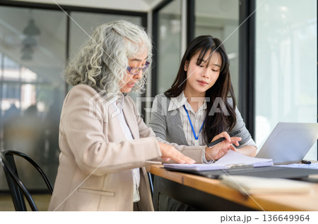 Old asian businesswoman holding pen explaining document to young coworker while sitting in office. 136649964