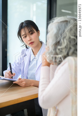Asian woman doctor writing pen on clipboard looking at old patient sits at table in diagnosis room 136649971