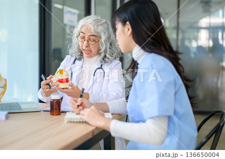 Old asian woman senior dentist holding tooth model teaching young doctor taking a note at exam table Old asian woman senior dentist holding tooth model teaching young doctor taking a note at exam table 136650004