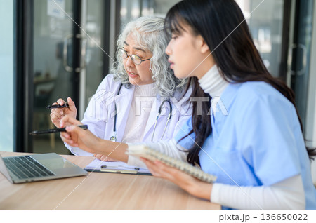 Old asian woman physician pointing on laptop showing to young doctor taking note sits at exam table. 136650022