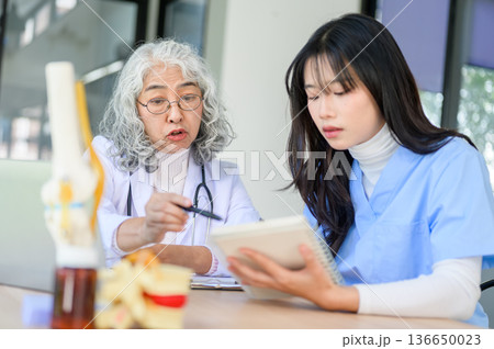 Old asian woman physician teaching and pointing at notebook of a young doctor sitting at exam table. Old asian woman physician teaching and pointing at notebook of a young doctor sitting at exam table. 136650023