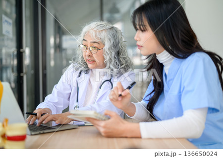 Old asian woman physician teaching and typing on laptop with young doctor taking note at exam table. Old asian woman physician teaching and typing on laptop with young doctor taking note at exam table. 136650024