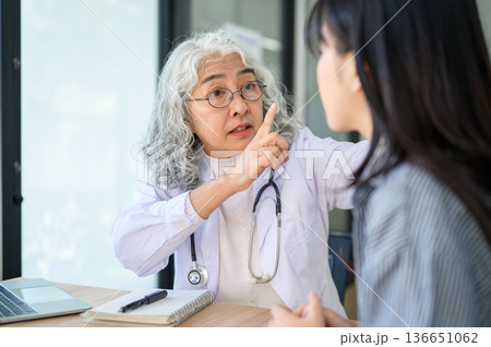 Old asian woman doctor warning or talking to young patient while sitting at table in diagnosis room. 136651062