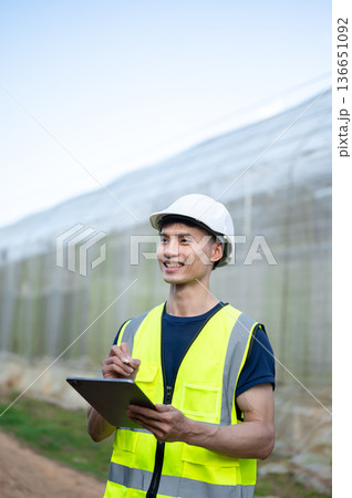 Asian engineer man wearing safety vest and hard hat helmet holding tablet standing aside greenhouse. Asian engineer man wearing safety vest and hard hat helmet holding tablet standing aside greenhouse. 136651092