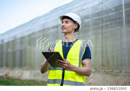 Asian engineer man wearings safety vest and hard hat helmet holding tablet standing aside greenhouse Asian engineer man wearings safety vest and hard hat helmet holding tablet standing aside greenhouse 136651093