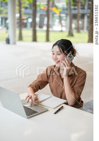 Asian woman student freelancer talking on phone and typing on laptop as sitting at table in building 136651382
