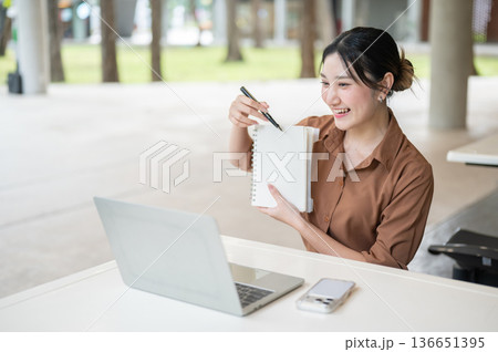 Asian woman student holding pen pointing on notebook across laptop at table in open air building. 136651395