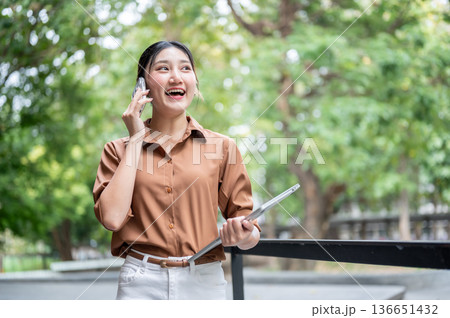 Asian woman student carrying laptop and talking on phone call while standing or walking in the park. Asian woman student carrying laptop and talking on phone call while standing or walking in the park. 136651432