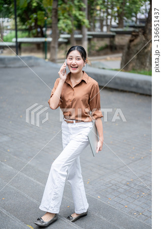 Smiling asian woman student talking on phone call carrying a laptop while walking on stairs in park. Smiling asian woman student talking on phone call carrying a laptop while walking on stairs in park. 136651437