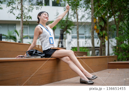 Asian office worker woman stretching arms as sitting cross legged with bag on wooden stairs in park. Asian office worker woman stretching arms as sitting cross legged with bag on wooden stairs in park. 136651455