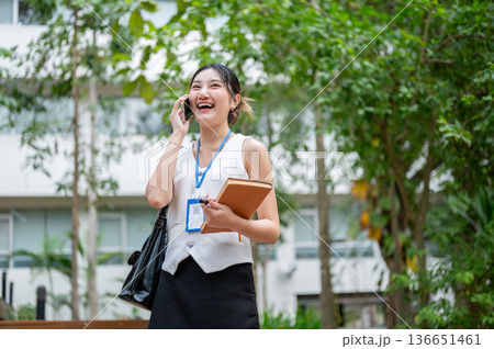 Asian woman office worker holding book talking on phone while walking or standing in a garden park. 136651461
