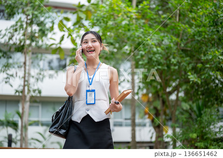Asian woman office worker carrying bag holding book talking on phone while walking standing in park. 136651462