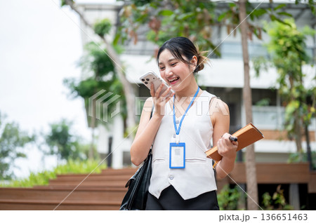 Asian woman office worker holding book talking on phone call while walking or standing in the park. 136651463