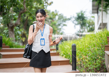 Asian woman office worker holding book talking on phone call while walking on wooden stairs in park. 136651472