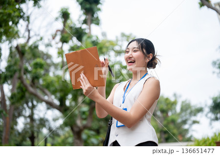 Happy asian woman office worker holding pen and looking at notebook while standing walking in park. 136651477