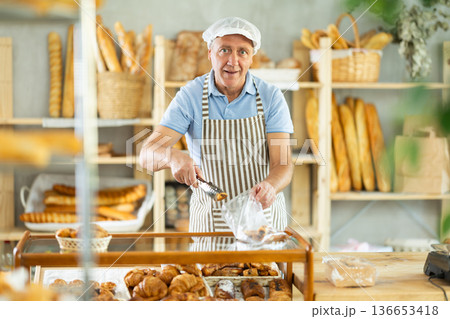 Senior man seller puts croissant in plastic disposable bag for buyer, packs goods. Senior man seller puts croissant in plastic disposable bag for buyer, packs goods. 136653418