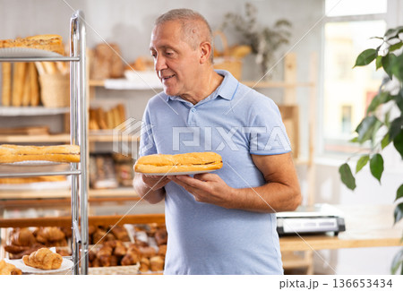 Senior male customer stands with croissant in hands near window of bakery. 136653434